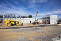 two construction workers stand outside an unfinished building under construction in a dirt area, with a crane to the side of the structure