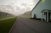 a beautiful house in the countryside during the day with fog in the air, a driveway leading to a barn with a white fence