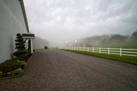 a beautiful house in the countryside during the day with fog in the air, a driveway leading to a barn with a white fence