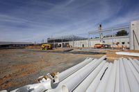 a dog sits on the ground next to pipes and a construction site in background, a man working