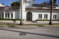 a white house with a red tiled roof and palm trees next to the road in front of it