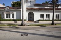 a white house with a red tiled roof and palm trees next to the road in front of it