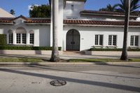 a white house with a red tiled roof and palm trees next to the road in front of it