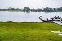 green grassy field next to wooden dock on the water shore with boats in the distance