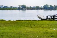 green grassy field next to wooden dock on the water shore with boats in the distance