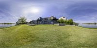 a large home surrounded by green grass and water with a blue sky and sun in the background