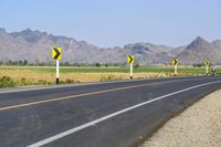 three yellow and white signs near a highway with mountains in the background in front of them