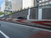 a black car drives on the highway below an overpass with tall buildings and cars driving down it