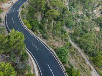 two cars traveling down an empty, winding, wide road between cliffs and trees and green vegetation