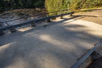 a concrete walkway leading to a river and two benches in the shade of trees next to it