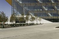 a skateboarder leaning against an asphalt parking lot near a tall glass building and trees