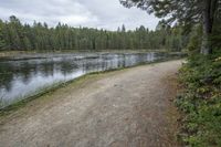 a man with a backpack walks across a gravel pathway beside a body of water near many trees