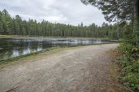 a man with a backpack walks across a gravel pathway beside a body of water near many trees