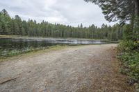 a man with a backpack walks across a gravel pathway beside a body of water near many trees