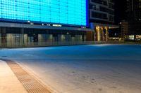 a blue and black lit up building next to a road at night time from the sidewalk