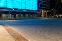 a blue and black lit up building next to a road at night time from the sidewalk