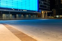 a blue and black lit up building next to a road at night time from the sidewalk