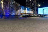 cars drive by in front of modern city buildings at night, including the lights on