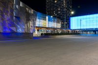 cars drive by in front of modern city buildings at night, including the lights on