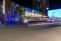 cars drive by in front of modern city buildings at night, including the lights on