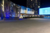 cars drive by in front of modern city buildings at night, including the lights on