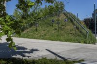 a green grassy hill and some trees on the side of a sidewalk near steps with arrows and signs in a circle