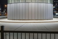 a photo taken at night of the circular building in an empty lot with a bench outside