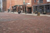 a large brick city street with people walking in it as well as tall buildings, with store fronts and buildings on the side and cars parked outside