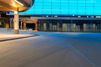 a paved street in front of a building with a blue glass wall above it at night