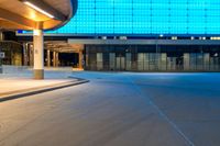 a paved street in front of a building with a blue glass wall above it at night