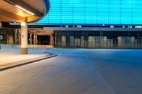 a paved street in front of a building with a blue glass wall above it at night