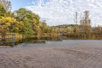 a brick walkway with benches near water and trees in the background against a bright blue sky