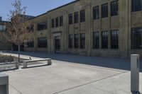 an empty courtyard with benches and tables in front of a brick building of a large, stone, factory or college building