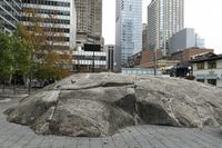 there is an artistic rock in the middle of a public square with tall buildings and skyscrapers