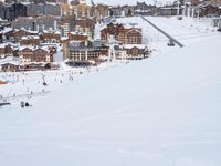 many people are skiing down a mountain near town buildings, snow covered slopes, and ski slopes with people skiing around in the distance