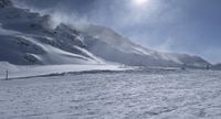 the snow is blowing with the clouds over the mountain side and snow slope in the foreground