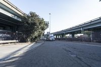 two trucks parked on the side of a street under a bridge near another bridge with a tall tree leaning over