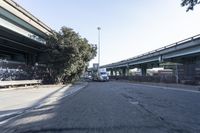 two trucks parked on the side of a street under a bridge near another bridge with a tall tree leaning over