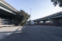 two trucks parked on the side of a street under a bridge near another bridge with a tall tree leaning over