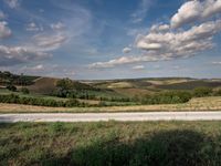 a country road running through a scenic landscape with rolling hills and trees around it and a cloud filled sky