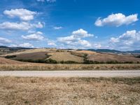 a field of land surrounded by a blue sky and many clouds in the distance a paved road leads to a hill