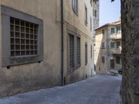 a narrow alley has buildings lining the street with windows and doors, including a small bench