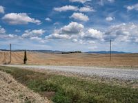 a dirt road passing by a field with a field in the distance and hills in the distance