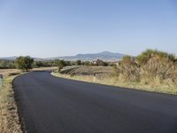 a rural road with no cars on the side of it near a grassy and mountains