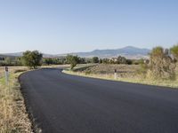 a rural road with no cars on the side of it near a grassy and mountains