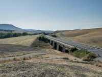 a train is traveling over a bridge on the road in rural country side with a valley and rolling fields on either side