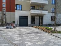 two bikes are parked against a row of residential buildings in a city driveway with brick pavers on the ground