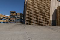 two men walk around with pallets stacked behind them in the sun next to a warehouse