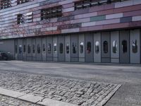 two people standing in front of a glass building on a brick walkway between two rows of doors