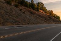 two people ride down the road on a motorcycle at sunset, passing by trees and hills